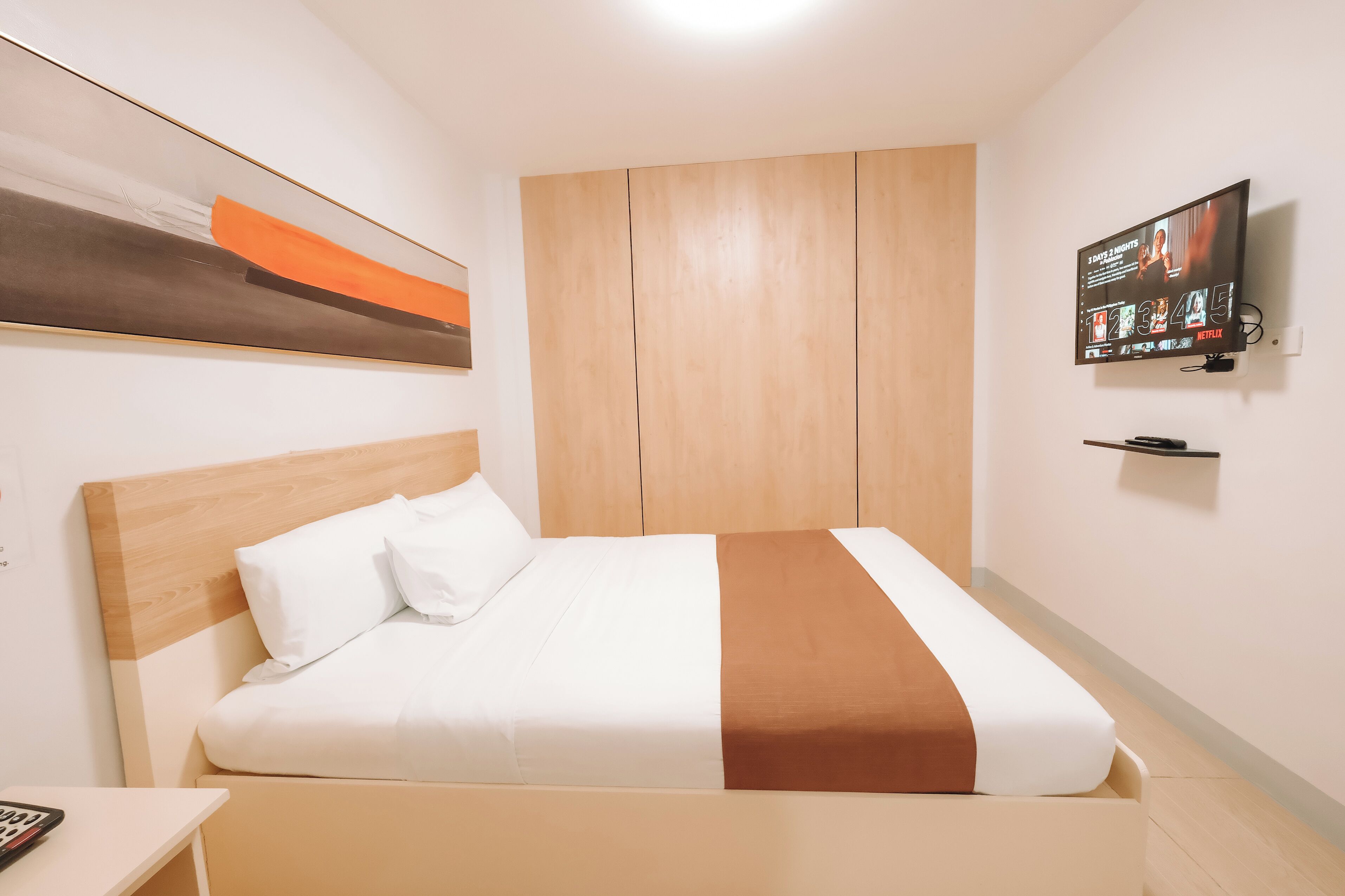 A hotel room with a bed, a TV mounted on the wall, and a wooden wardrobe.