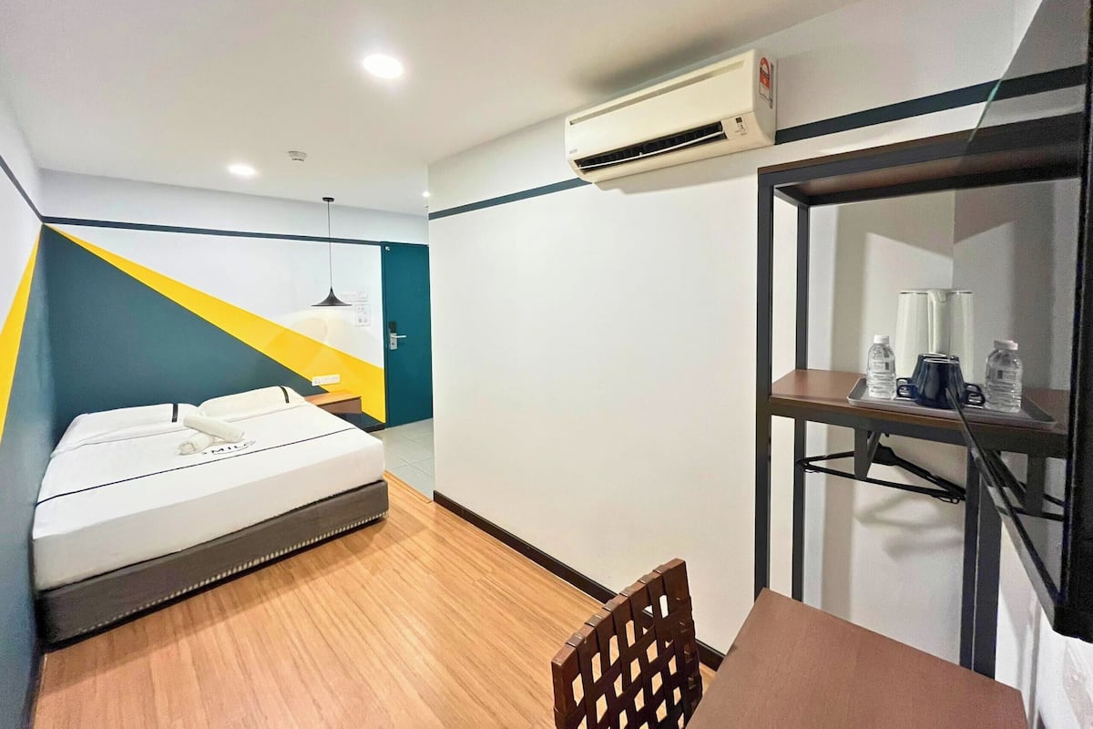 Une chambre d’hôtel avec un lit, un bureau et une chaise, un appareil de climatisation et une étagère avec des bouteilles.