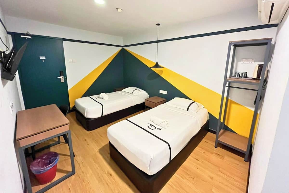 Une chambre d’hôtel avec deux lits simples, un petit bureau, une poubelle et une étagère avec des articles de toilette.