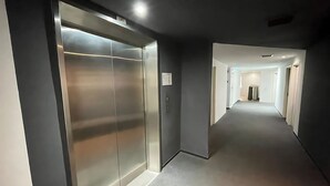 Ein moderner Aufzug mit Metalloptik und einem Schild, das die Etage angibt. Ein Flur mit grauem Teppich und weißen Wänden führt zu einer Tür am Ende.