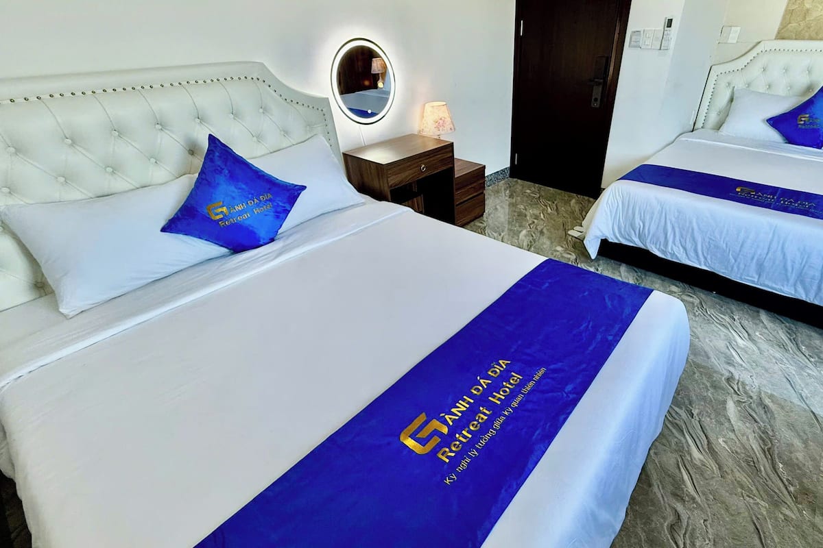 A hotel room with two beds, each with a blue bedspread and a white headboard. There is a nightstand with a lamp and a mirror between the beds.