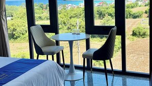Habitación de hotel con una cama grande, dos sillas, una mesita y vistas al mar y al follaje.