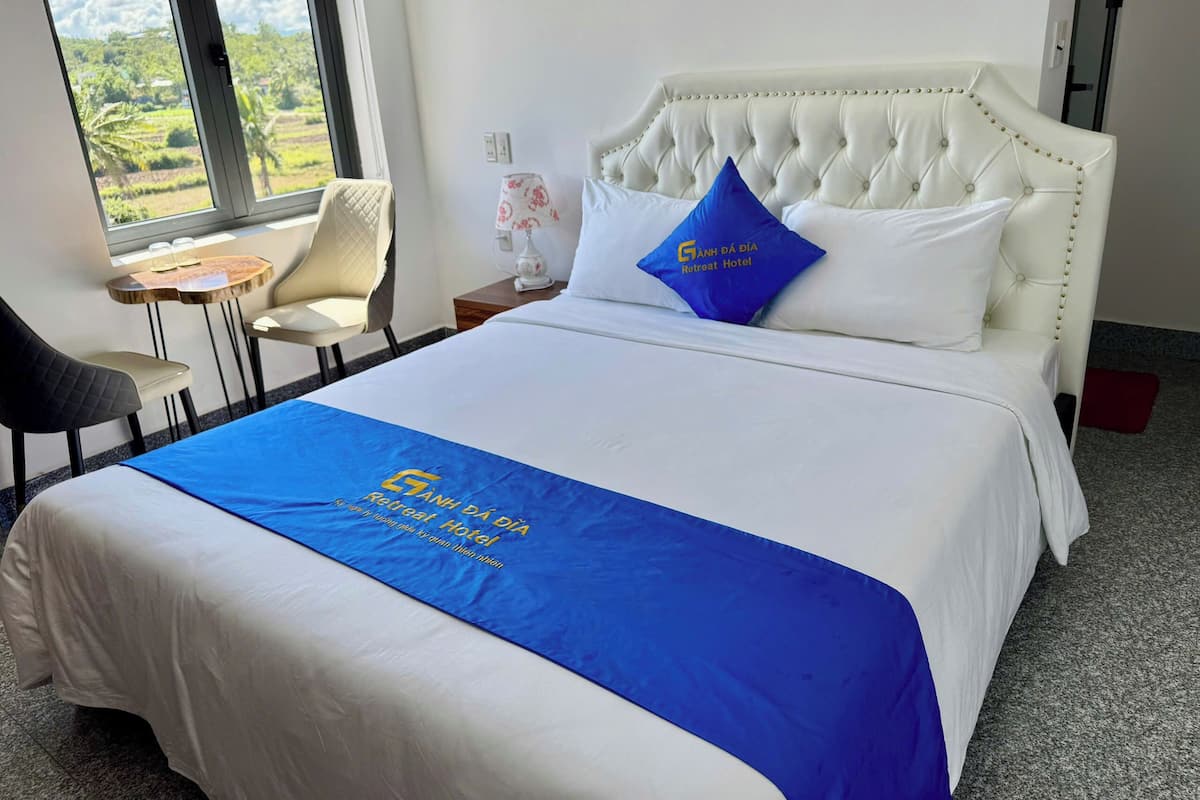 A hotel room with a bed, a window with a view of greenery, a small table with chairs, and a nightstand with a lamp.