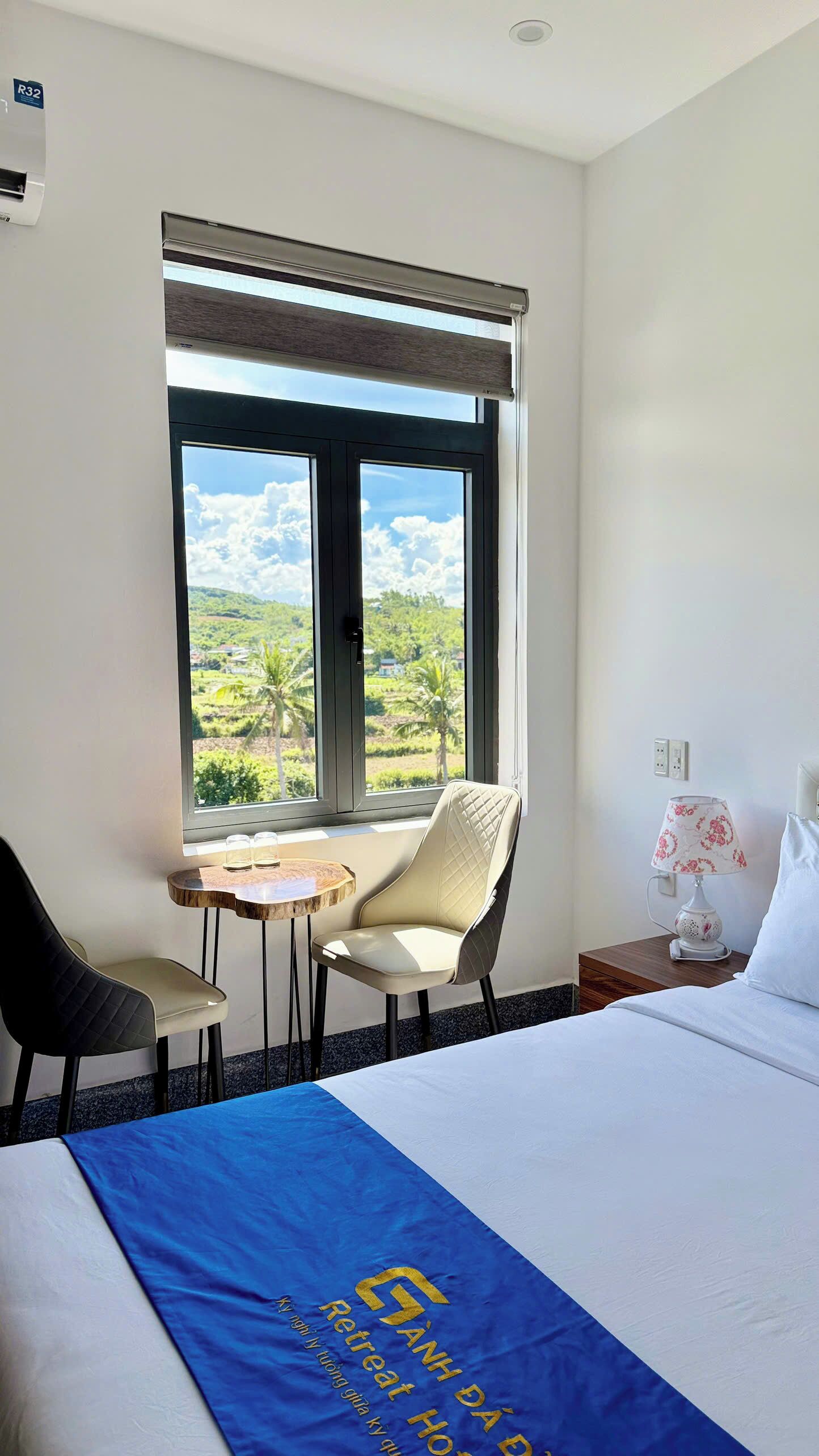 A hotel room with a bed, a window with a view of palm trees and a cloudy sky, a small table with chairs, and a lamp on a bedside table.