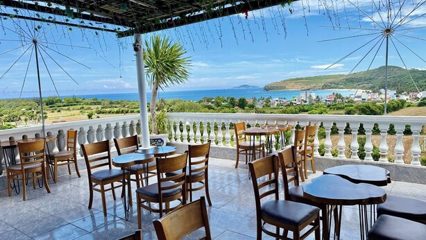 Una terraza con mesas y sillas, con vistas a un paisaje costero con vegetación y edificios.