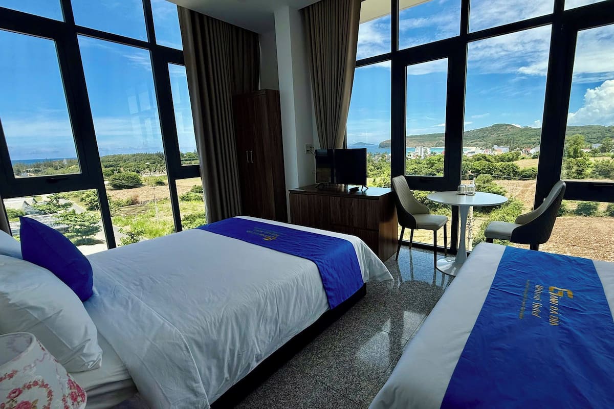A hotel room with two beds, a desk, a chair, and a small table. The room has large windows offering a view of the countryside.