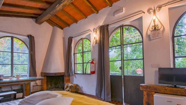 Una habitación con cama, escritorio, televisor y ventanas grandes con cortinas.