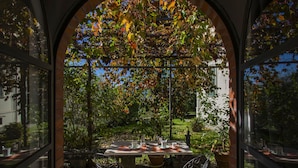 Un espacio de comedor al aire libre con una mesa dispuesta para dos personas, rodeado de un muro de ladrillo y ventanas grandes.