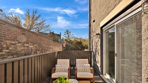 A balcony with two wooden chairs, a small table with a plant and decorative items, and a view of a brick wall and trees. - Vital Urban: Spa Inspired Retreat (Chicago)
