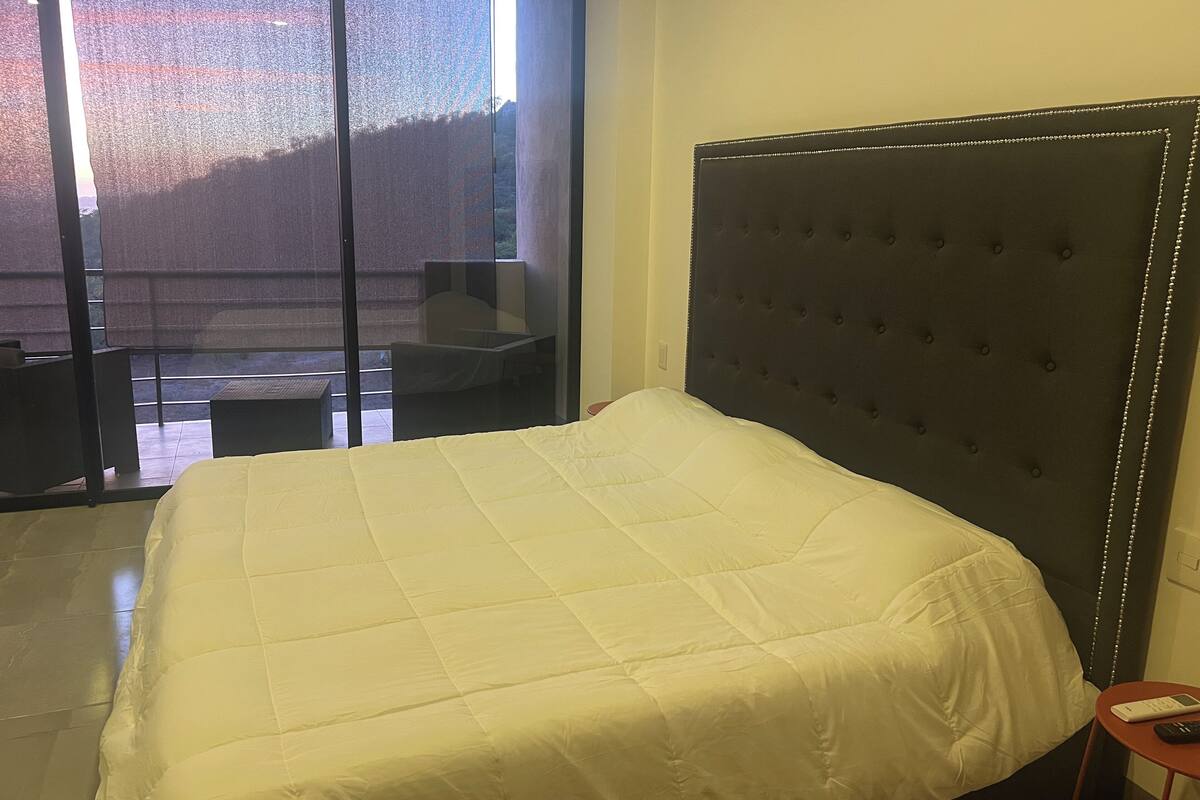 A hotel room with a large bed, a dark upholstered headboard, a small table with a remote control, and a view of a balcony with outdoor furniture.