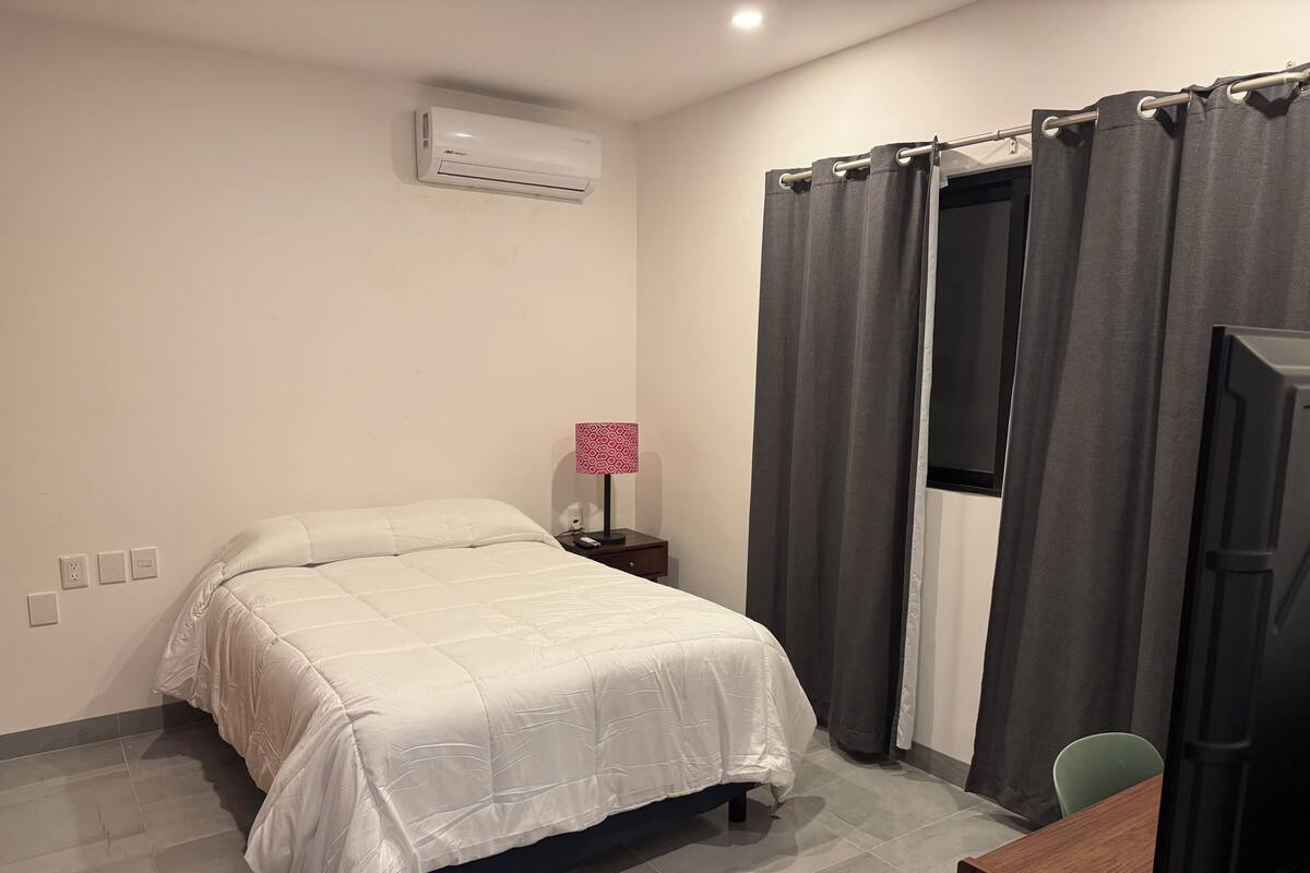 A hotel room with a bed, a desk, a chair, a lamp, and an air conditioning unit.