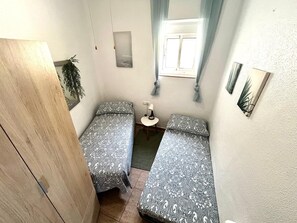 Una habitación pequeña y funcional con dos camas individuales, una mesita, una lámpara y una ventana con cortinas.