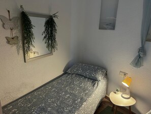 A small bedroom with a bed, a bedside table with a lamp, a mirror, and a wall-mounted plant.
