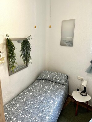 Een kleine, verzorgde slaapkamer met een eenpersoonsbed, een tafeltje, een lamp en een spiegel aan de muur met hangplanten.