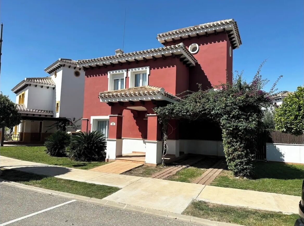 Une maison à deux étages avec une façade rouge et des ornements blancs, un porche à l’avant et un petit jardin.