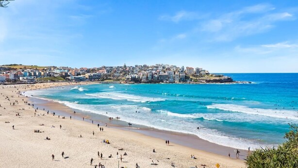A coastal town with a wide sandy beach, clear blue waters, and a densely built urban area in the background.