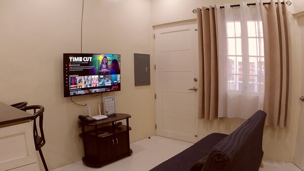 A room with a bed, a desk with a computer, a TV mounted on the wall, a door, and a window with curtains. - Entire home/Fully furnished/Netflix/AC/Wash/Dyer (Daet)