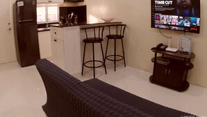 A compact kitchen with a refrigerator, sink, and bar stools. A flat-screen TV mounted on the wall displays a movie. - Entire home/Fully furnished/Netflix/AC/Wash/Dyer (Daet)