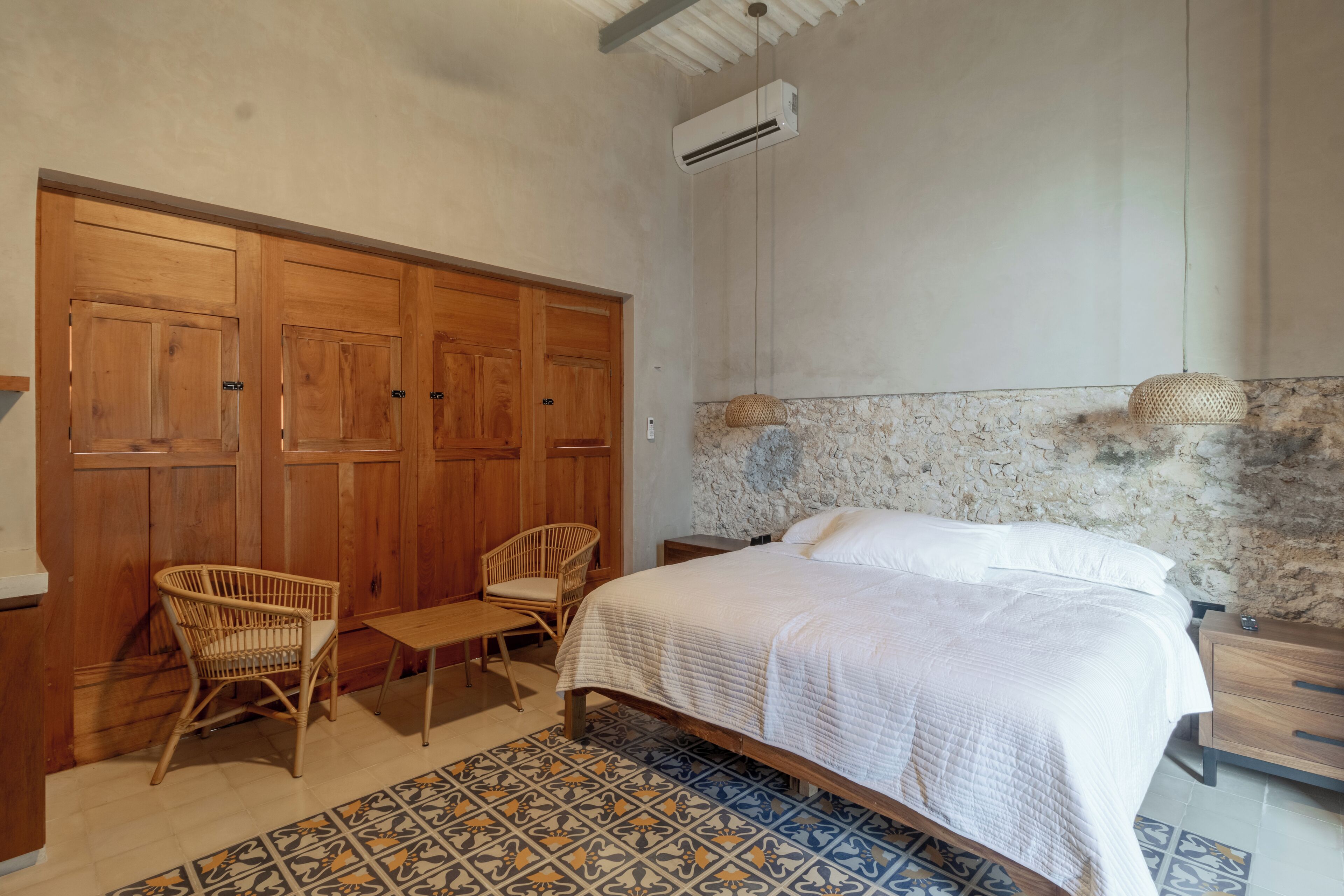 Quarto com cama, duas cadeiras de vime, uma mesinha, guarda-roupa de madeira e parede com padrão de pedra.