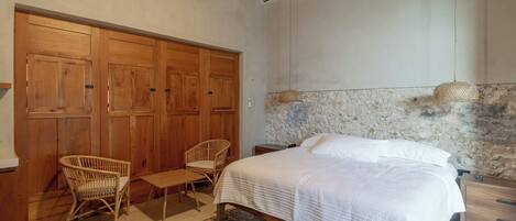 Quarto com cama, duas cadeiras de vime, uma mesinha, guarda-roupa de madeira e parede com padrão de pedra.