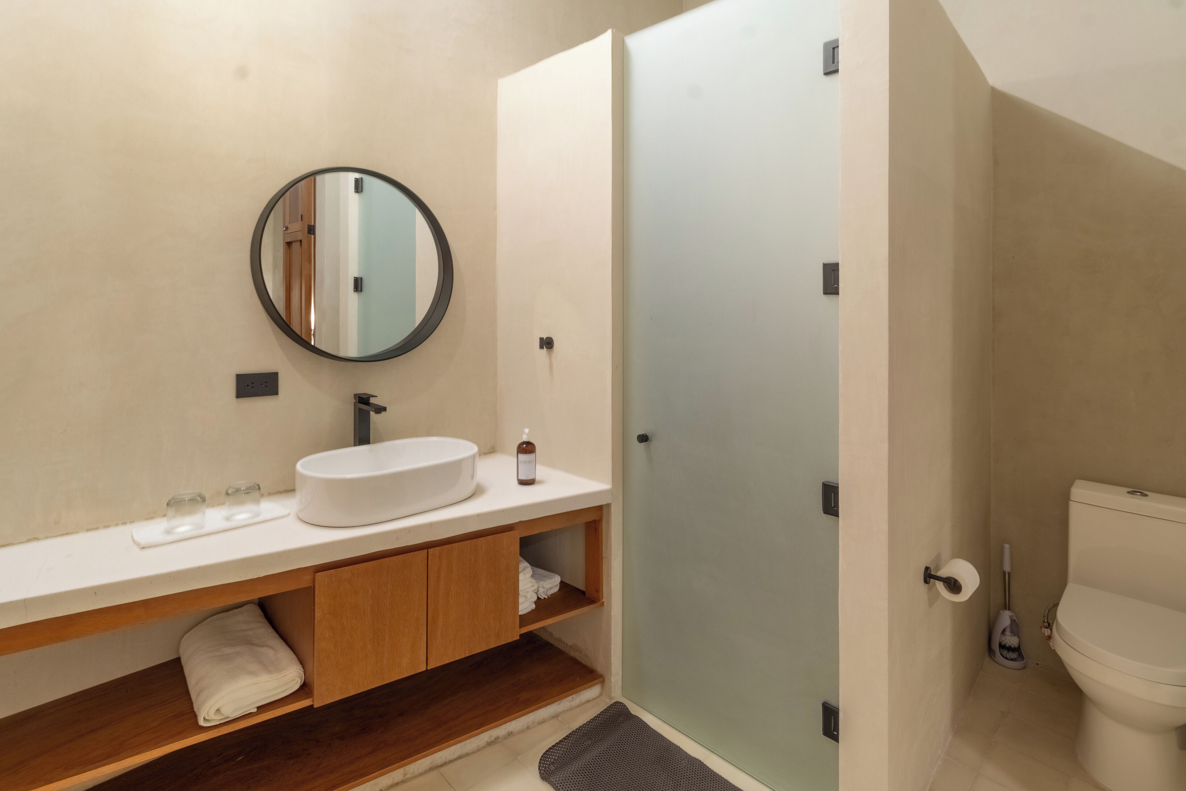 A modern bathroom with a round mirror, a white sink, wooden cabinets, and a glass shower enclosure.