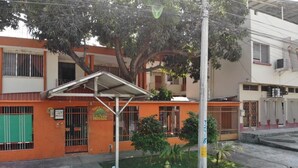 Un edificio de dos plantas con un área de estar al aire libre cubierta, un pequeño jardín y una entrada visible con una reja de metal.