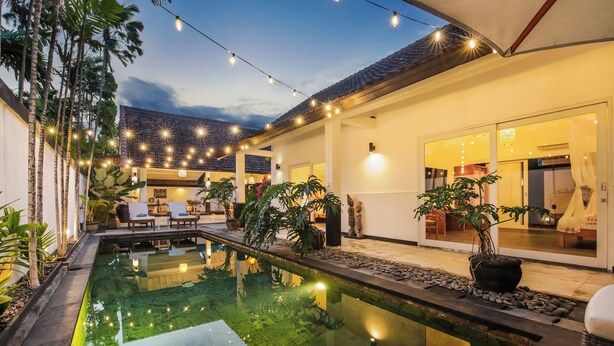 A pool area with string lights, a patio with chairs, and a view of the interior. - Villa Zebra (Seminyak)