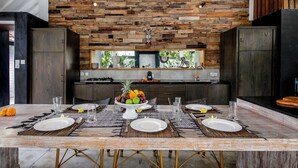 A dining area with a rustic wooden wall, a kitchen in the background, and a fruit centerpiece on the table. - Villa Zebra (Seminyak)