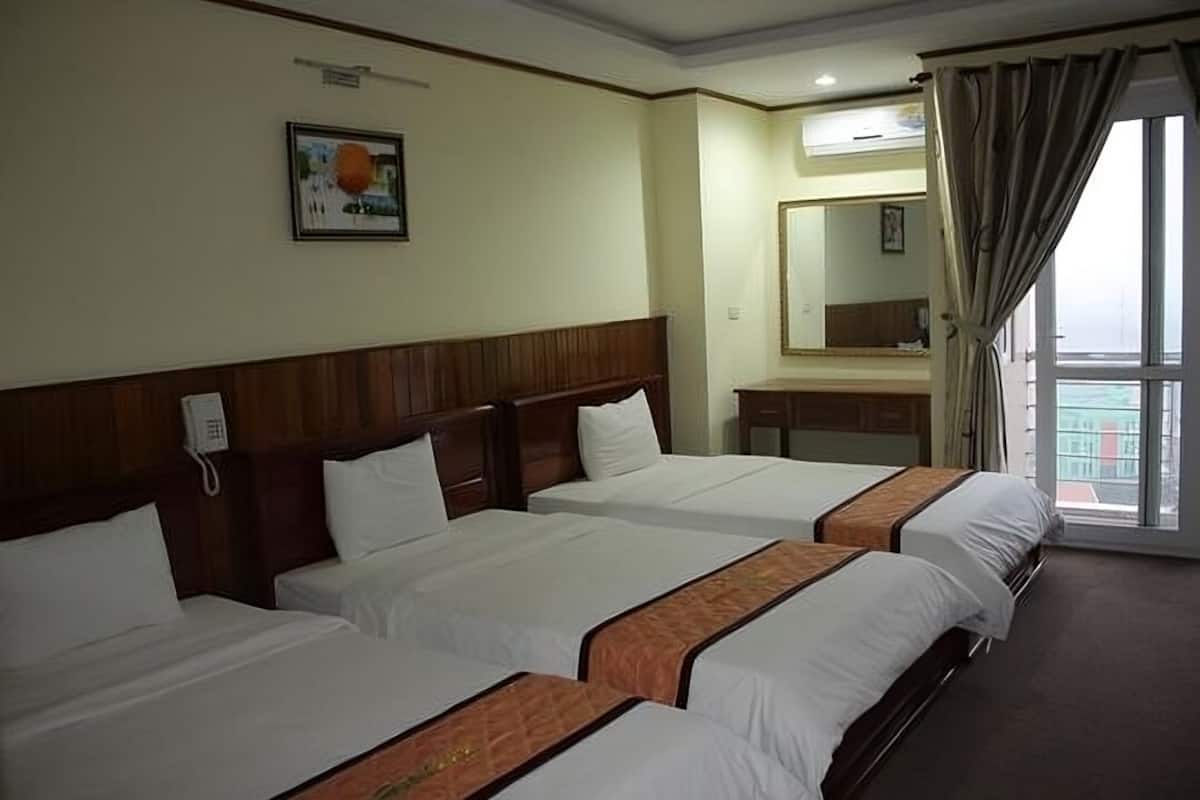 A hotel room with three beds, a wooden headboard, a telephone, a painting on the wall, a large window with curtains, and a bathroom with a mirror.