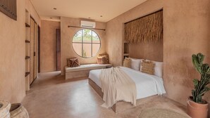 Een slaapkamer met een groot bed, een rond raam, een kamerplant en een luchtverkoeler aan de muur.