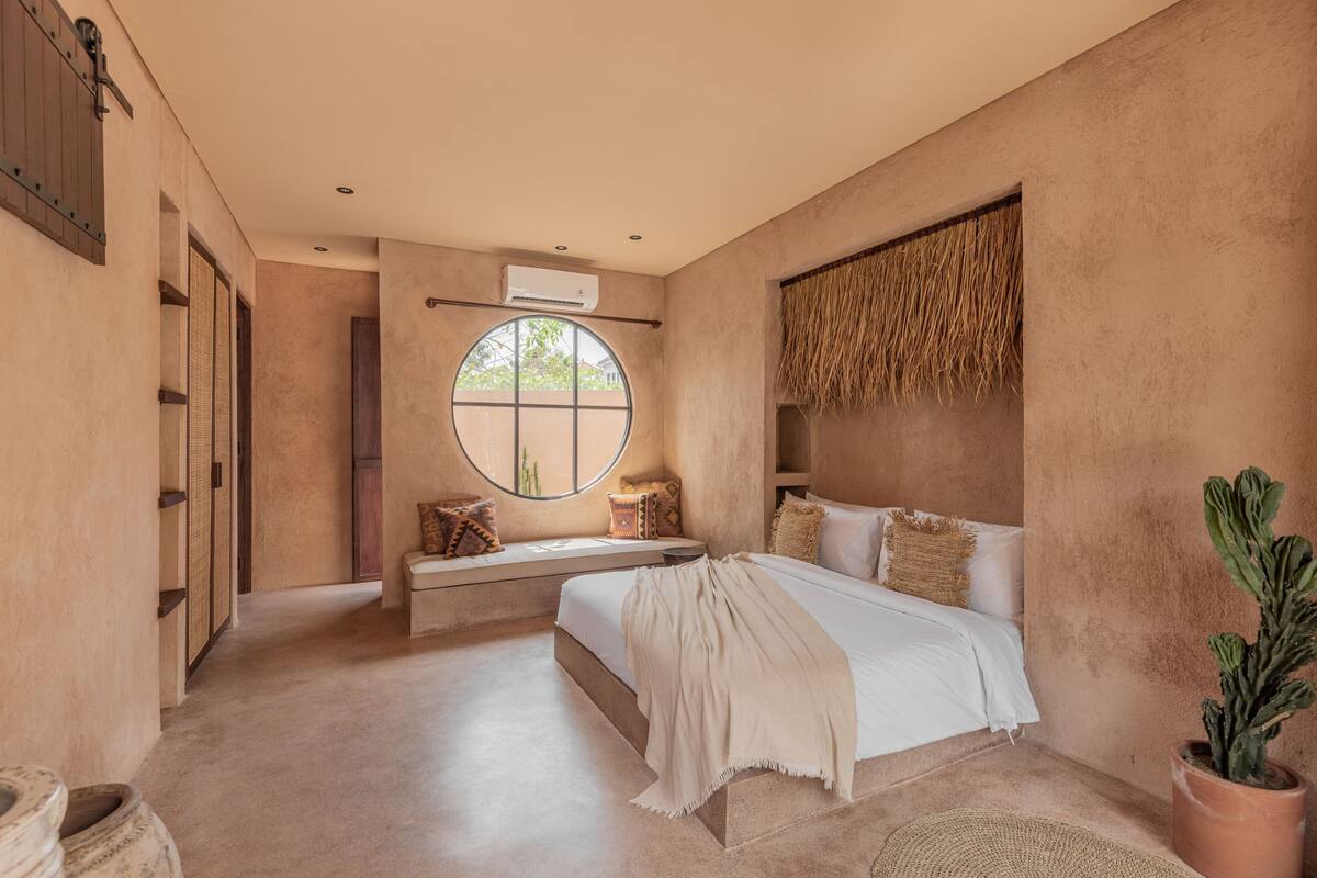 A bedroom with a large bed, a round window, a potted plant, and a wall-mounted air conditioner.