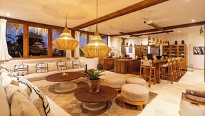 A spacious living room with a large sofa, round coffee table, and ottomans. There are hanging pendant lights and a ceiling fan. A kitchen area with wooden cabinets and bar stools is visible in the background.