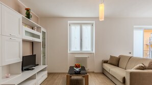 Een moderne woonkamer met een houten vloer, een beige bank, een tv op een witte kast en een raam met uitzicht.