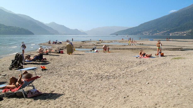 Een strand met mensen die zonnebaden en zwemmen, omringd door bergen en een heldere lucht.