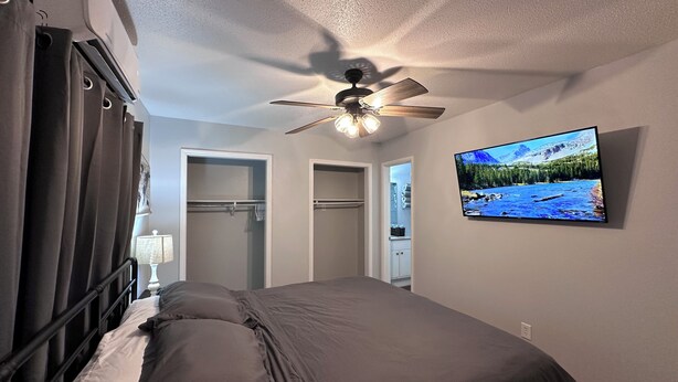 Ein Schlafzimmer mit Bett, Deckenventilator, Fernseher an der Wand und Kleiderschrank mit Spiegel.