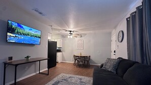 Ein Wohnzimmer mit Flachbildfernseher, Deckenventilator, Essbereich mit Tisch und Stühlen sowie einem dunkelgrauen Sofa.