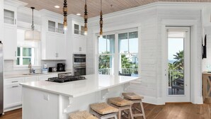 A modern kitchen with white cabinetry, a stainless steel refrigerator, a built-in oven, and a large island with bar stools. - Sea You Later by Kaiser Vacation Rentals Cottages at Romar 15 (Orange Beach)