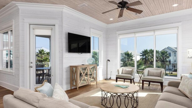 A spacious living room with a ceiling fan, a flat-screen TV, and a large window offering a view of palm trees and a house. - Sea You Later by Kaiser Vacation Rentals Cottages at Romar 15 (Orange Beach)