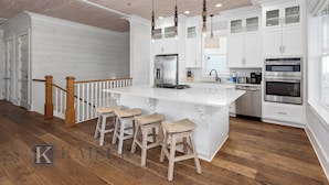 A modern kitchen with white cabinetry, a large island with bar stools, stainless steel appliances, and a wooden staircase. - Sea You Later by Kaiser Vacation Rentals Cottages at Romar 15 (Orange Beach)