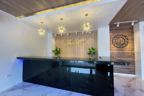 Un área de recepción con mostrador de mármol negro, candelabros modernos, una pared de madera con un reloj y dos plantas en macetas.