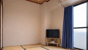 Una habitación con suelo de tatami, un televisor sobre un soporte de madera, un aire acondicionado y una ventana con cortinas.