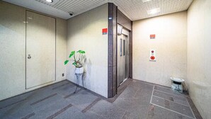 Un área de ascensor limpia y moderna con suelo de baldosas, una planta en maceta y un pequeño cubo de basura.