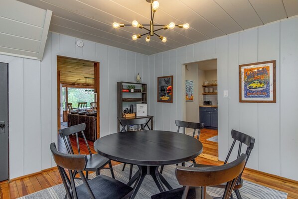 A dining area with a round table and chairs, a kitchen in the background, and a ceiling light fixture.