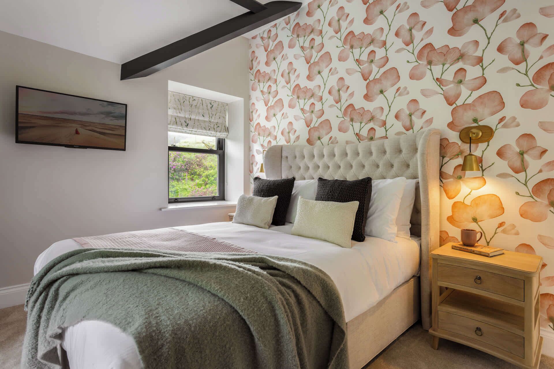 Un dormitorio con una cama grande, una mesita de noche, una ventana que da a vistas de vegetación y una pared con estampado floral.