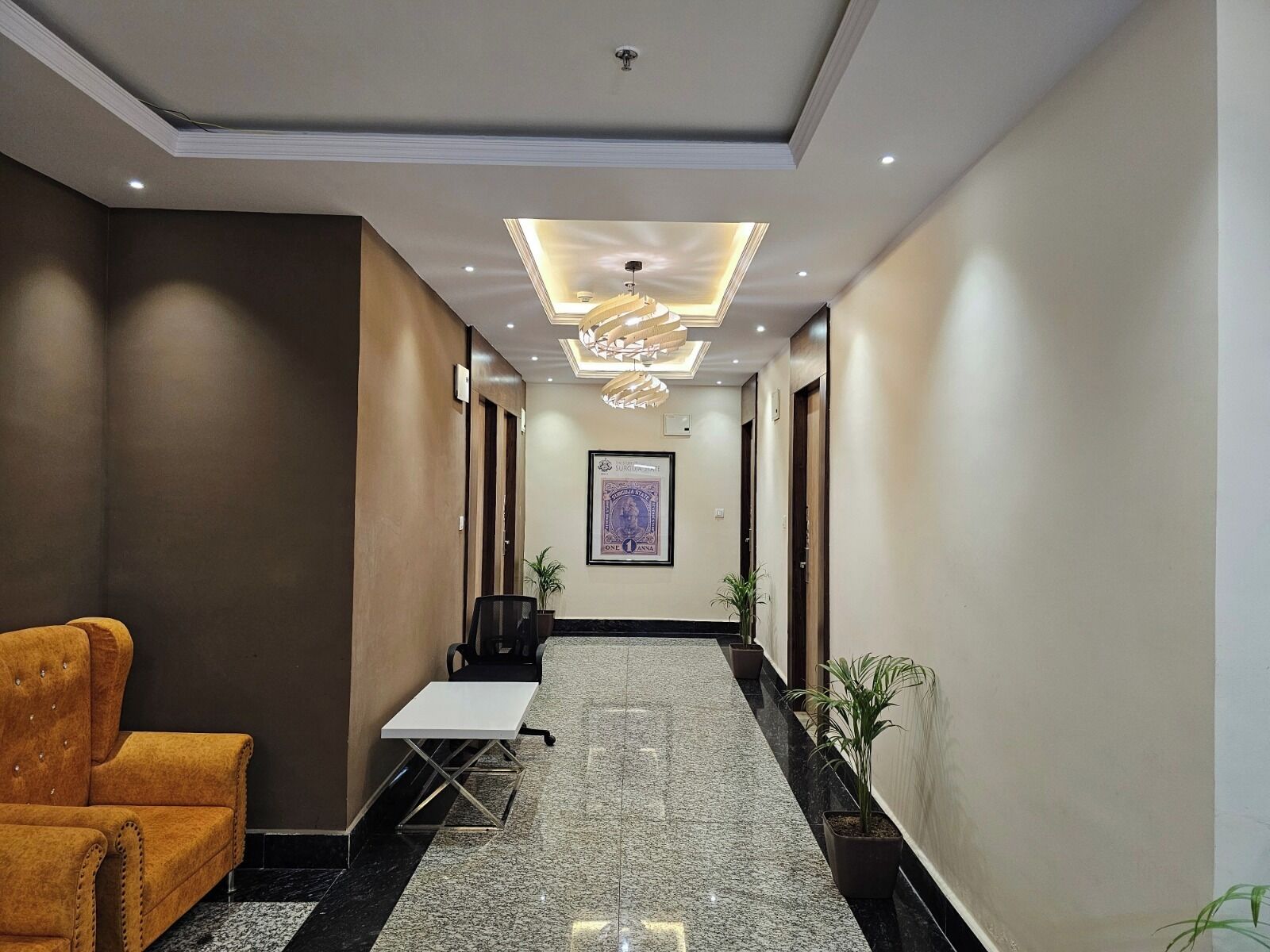 Un couloir d’hôtel agrémenté d’un lustre moderne, d’une affiche encadrée au mur, d’un coin salon avec un canapé et de plantes en pot disposées le long du couloir.