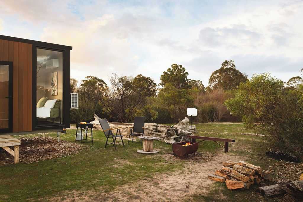 Une cabane moderne dotée d’une porte en verre, d’un foyer extérieur, de sièges pour se détendre en plein air et offrant une vue sur la forêt environnante.