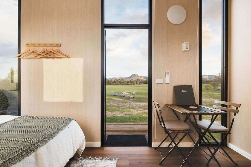 Een slaapkamer met een bed, een bureau met een laptop en uitzicht op een grasachtig landschap door een groot raam.