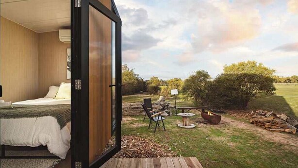 A bedroom with a large window, a bed, and a view of an outdoor patio with a fire pit and seating area. - Hollow Mountain Getaway 1 by Tiny Away (Laharum)