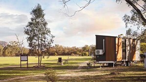 A modern RV with a wooden exterior parked in a grassy area with trees and a signboard in the background.