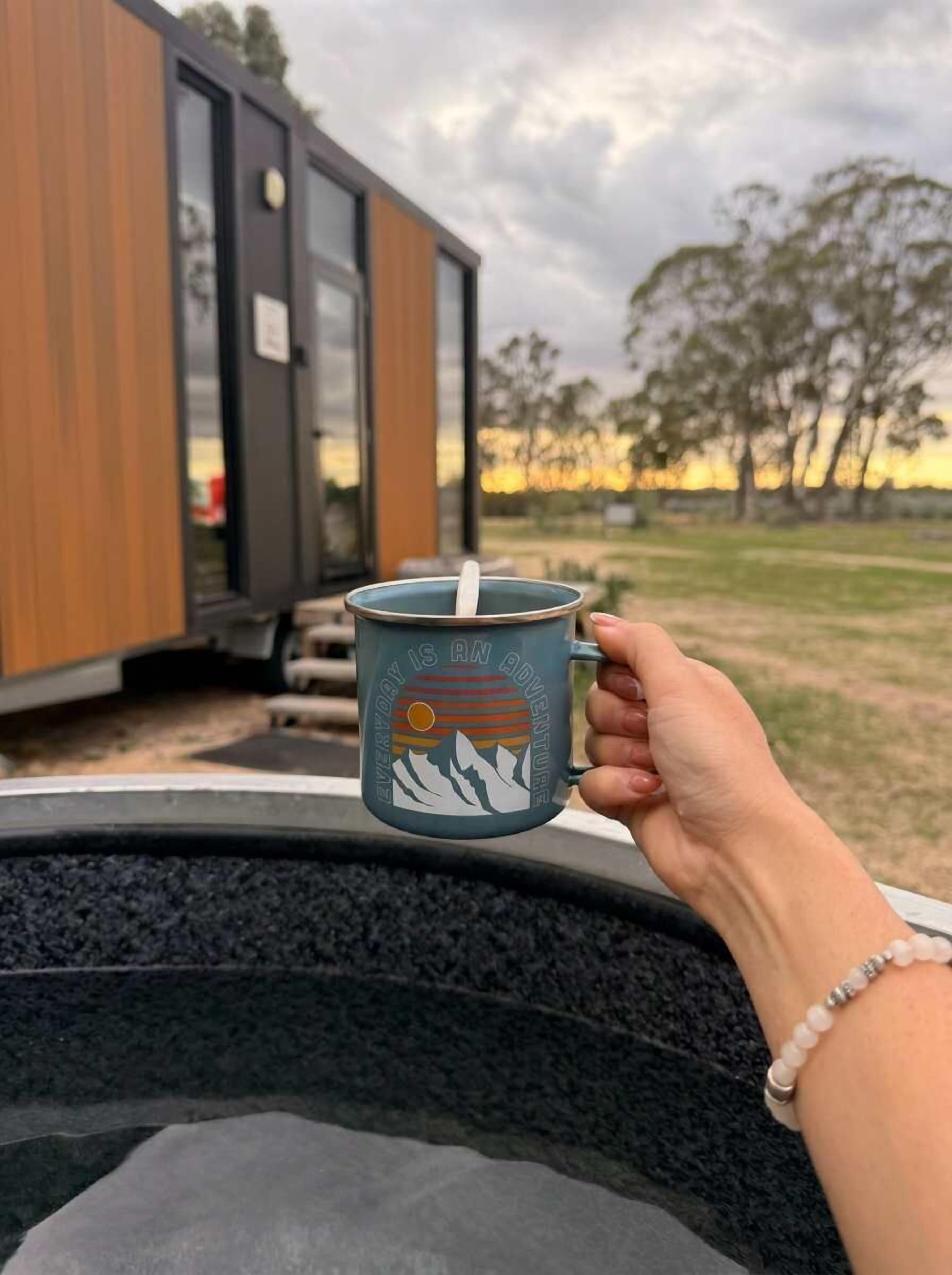 Une main tenant une tasse ornée d’un motif représentant une montagne et un soleil, posée devant un bâtiment moderne doté de grandes fenêtres.